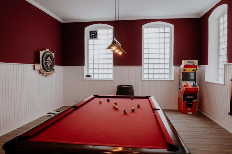 Roter Billiardtisch vor dunkler Wand mit Dartscheibe und Spielautomaten